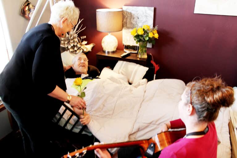 woman playing a guitar for an elderly man in a hospice bed and a visitor